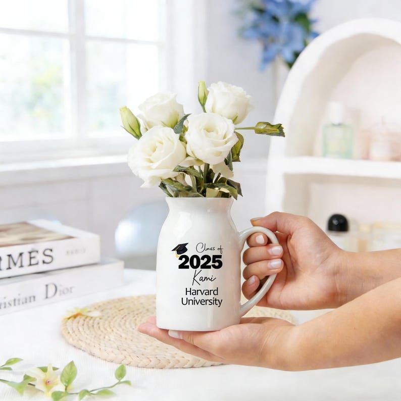 May include: A white ceramic vase with a handle, filled with white roses. The vase has black text that reads "Class of 2025 Kami Harvard University". The vase is held in two hands.