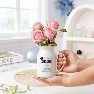 May include: A white ceramic vase with a handle, filled with pink roses. The vase has the text "Class of 2025 Kami Harvard University" printed in black. The vase is held in someone's hands.