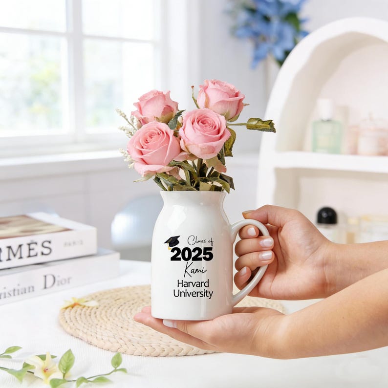 May include: A white ceramic vase with a handle, filled with pink roses. The vase has the text "Class of 2025 Kami Harvard University" printed in black. The vase is held in someone's hands.