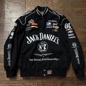 Puede incluir: Chaqueta de carreras negra con el logotipo de Jack Daniel's y parches de patrocinadores. La chaqueta tiene cierre de botones a presión, cuello alzado y el texto "Pace Yourself. Drink Responsibly." en la parte delantera.