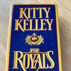 Könnte beinhalten: Ein blaues Hardcover-Buch mit gelbem Rücken und einem goldenen Kronendesign. Der Titel des Buches lautet "Kitty Kelley The Royals".