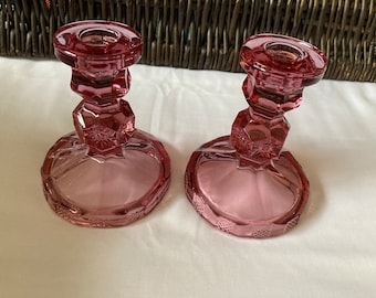 May include: Two pink glass candle holders with a geometric design. The holders are made of clear glass with a pink tint.