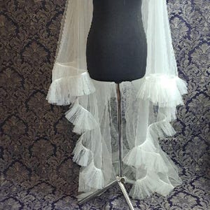 May include: A white tulle bridal veil with multiple layers of ruffles, displayed on a black mannequin. The veil is long and flowing, with a delicate, airy appearance. The background features a patterned fabric.