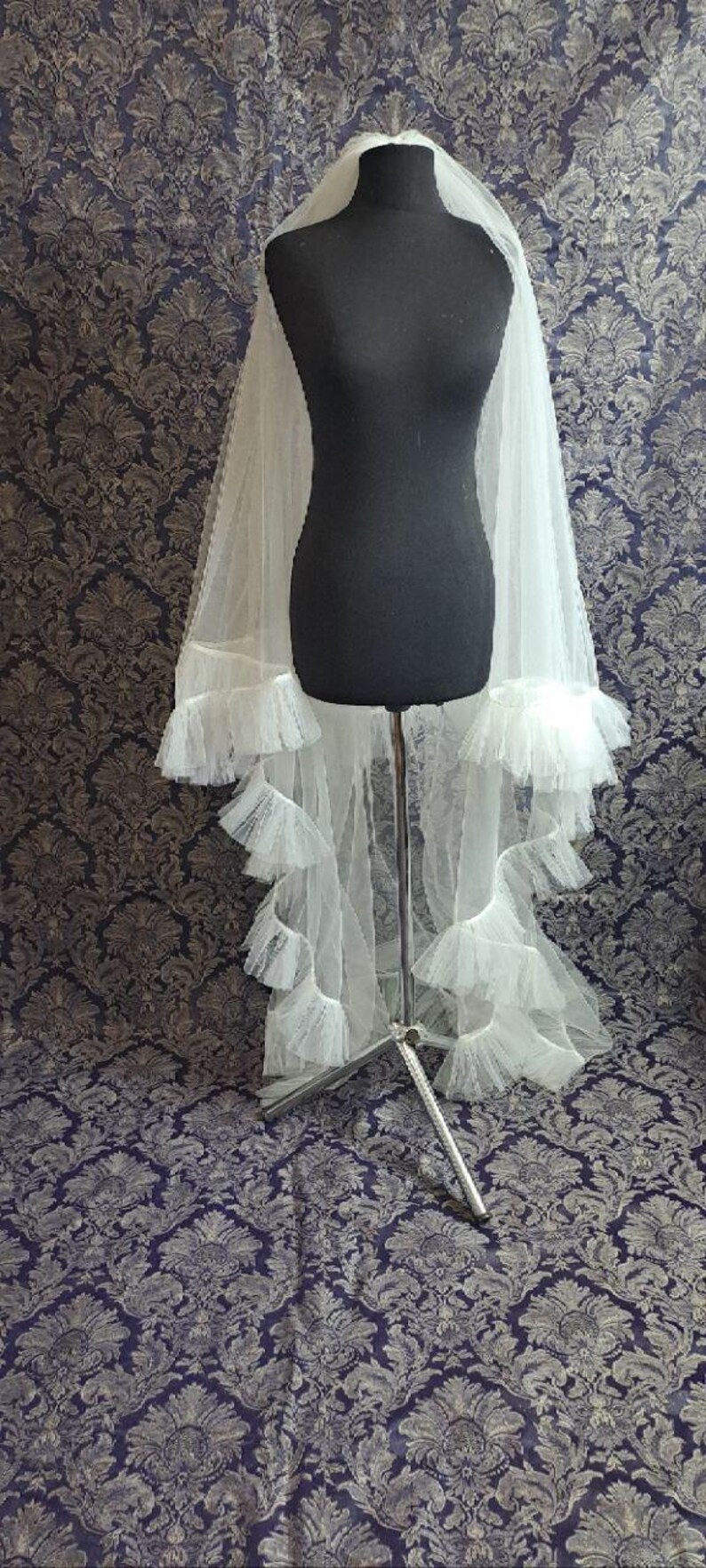 May include: A white tulle bridal veil with multiple layers of ruffles, displayed on a black mannequin. The veil is long and flowing, with a delicate, airy appearance. The background features a patterned fabric.