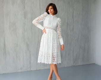 May include: A white lace dress with long sleeves and a high neckline. The dress features a fitted bodice and a flared skirt that falls to the mid-calf. The dress is made of delicate lace and has a feminine and elegant design.
