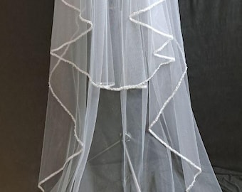 May include: A white bridal veil with multiple layers and a decorative, beaded edge. The veil is made of a sheer fabric and hangs vertically, showcasing its flowing design. The veil is a classic wedding accessory.