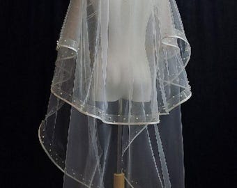 May include: A sheer white bridal veil with a double-layered design. The veil is adorned with small, evenly spaced pearls along the edges and is displayed on a mannequin. The background is a dark, solid color.