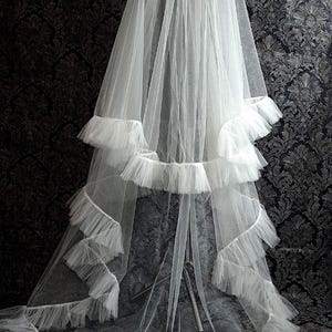 May include: A white bridal veil with multiple layers of delicate tulle and ruffled edges. The veil is long and flowing, with a tiered design and a soft, ethereal appearance. The background features a dark, patterned fabric.