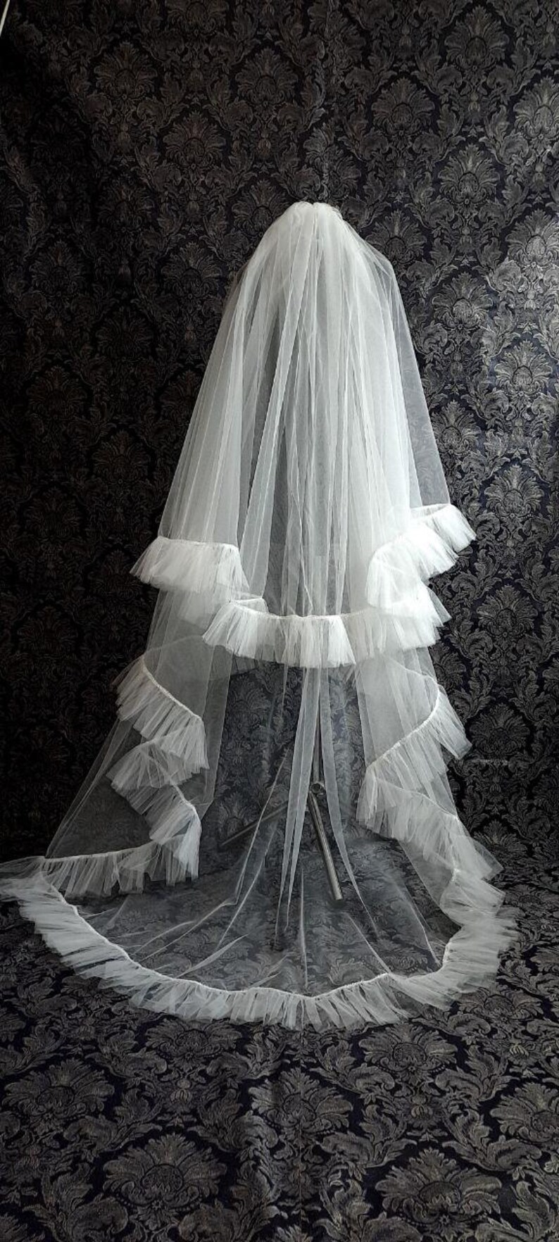 May include: A white bridal veil with multiple layers of delicate tulle and ruffled edges. The veil is long and flowing, with a tiered design and a soft, ethereal appearance. The background features a dark, patterned fabric.