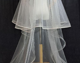 May include: A two-tiered, white bridal veil with a delicate, sheer fabric. The veil is displayed on a mannequin, showcasing its flowing design. The edges are trimmed with a thin, white satin ribbon.
