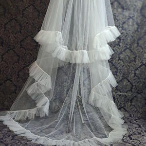 May include: A white bridal veil with multiple layers of sheer fabric and ruffled trim. The veil is displayed against a patterned background, showcasing its delicate texture and design. A classic accessory for weddings.