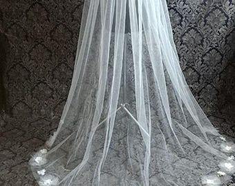 May include: A long, white bridal veil with a delicate floral trim. The veil is made of sheer fabric and is displayed on a stand against a patterned backdrop. The veil's length is visible, and the floral trim adds a touch of elegance.