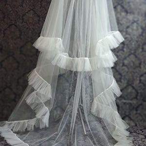 May include: A white bridal veil with multiple layers of soft tulle. The veil features a tiered design with ruffled edges, creating a flowing and elegant look. The veil is displayed against a patterned background.