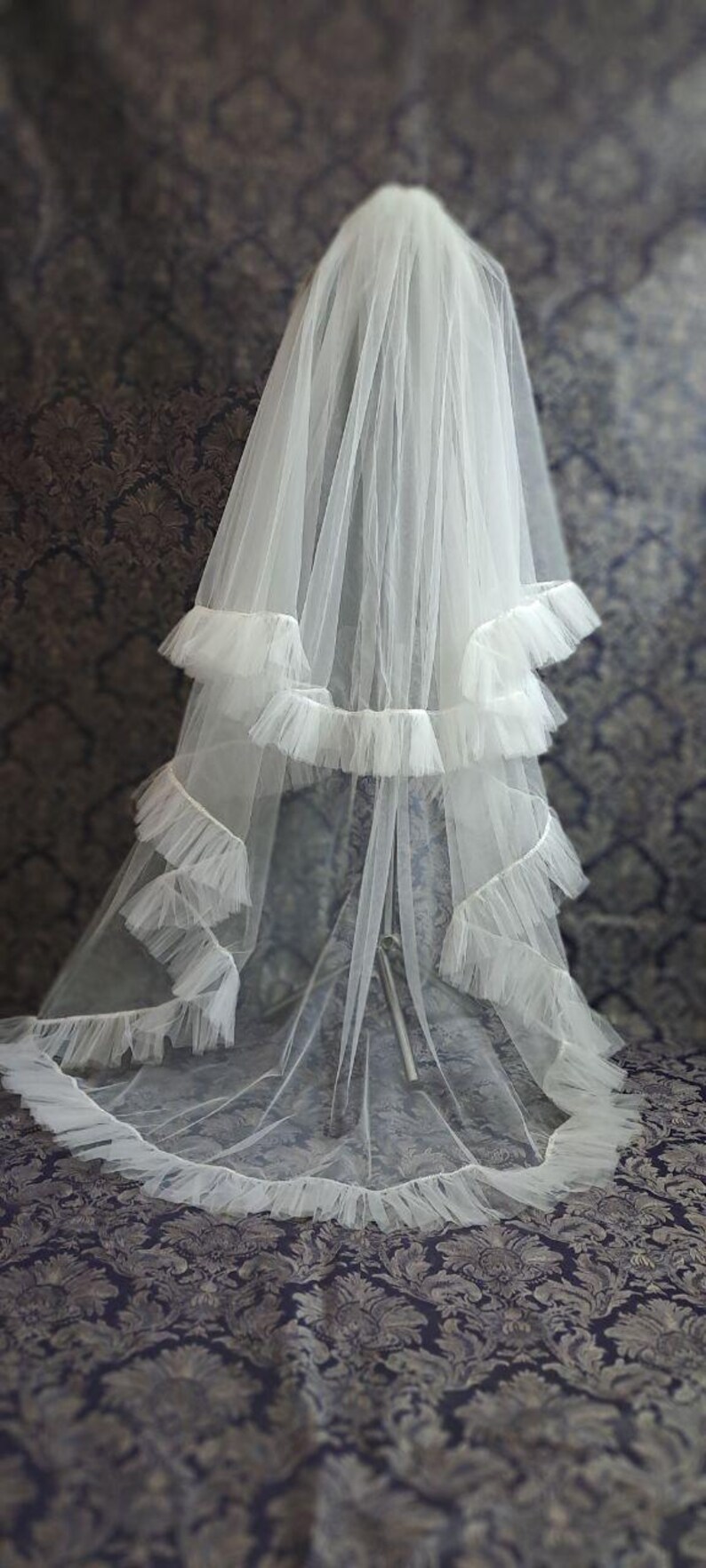 May include: A white bridal veil with multiple layers of soft tulle. The veil features a tiered design with ruffled edges, creating a flowing and elegant look. The veil is displayed against a patterned background.