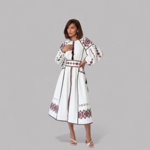 May include: White midi dress with intricate embroidery in red and black. The dress features long, puffy sleeves, a high neckline, and a fitted waist. The embroidery is concentrated on the sleeves, bodice, and hemline, creating a folk-inspired aesthetic. The dress is paired with white sandals.