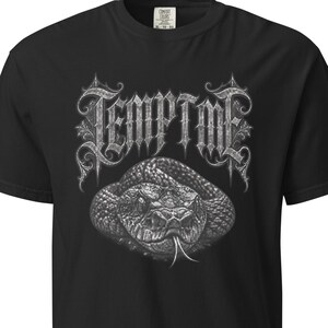 Op de afbeelding: Een zwart t-shirt met korte mouwen en een grafische print van een opgerolde slang met een gespleten tong. Boven de slang staat het woord TEMPTME in een gestileerd, sierlijk, metaalgrijs lettertype met ingewikkelde details.