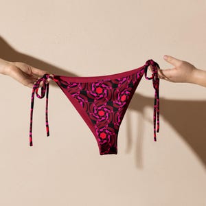 Peut inclure: Bas de bikini bordeaux avec un motif géométrique rose et noir. Le bas de bikini a des liens sur les côtés. Le maillot de bain est tenu par deux mains.