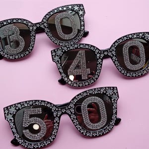 May include: Three pairs of black sunglasses with rhinestone numbers 30, 40, and 50. The frames are decorated with rhinestones and star-shaped accents. The lenses are mirrored, set against a pink background.