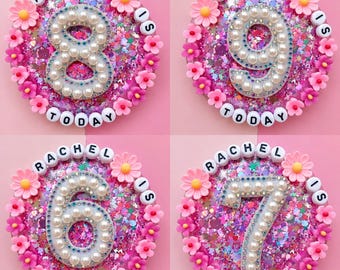 May include: Four circular resin plaques filled with pink glitter and confetti. Each plaque features a large number outlined in white pearls and blue rhinestones, surrounded by pink and yellow flower charms and white letter beads spelling RACHEL IS TODAY.