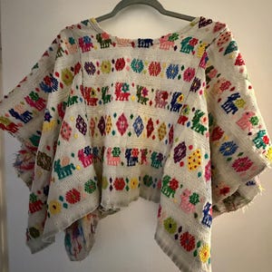Puede incluir: Una blusa blanca de manga corta y corte holgado con bandas horizontales de bordados coloridos e intrincados. Los patrones incluyen formas geométricas repetitivas, motivos florales y figuras animales estilizadas en tonos vibrantes de rojo, azul, verde, amarillo y rosa.