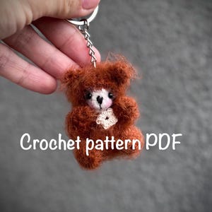 Puede incluir: Un pequeño llavero de oso de peluche tejido en color marrón, con un detalle de flor blanca. El oso tiene una textura esponjosa y está unido a un llavero plateado. El texto en la imagen dice "Crochet pattern PDF".