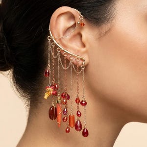 May include: Gold-toned ear cuff with dangling red and orange beads and charms. The ear cuff wraps around the ear and features multiple chains with various bead shapes and a small figurine. A stylish accessory.