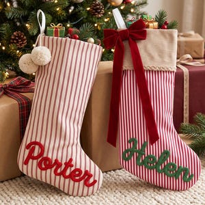 Puede incluir: Dos calcetines navideños con rayas verticales rojas y blancas. El calcetín izquierdo tiene dos pompones color crema y el nombre Porter en letra roja. El derecho tiene un gran lazo de terciopelo rojo y el nombre Helen en letra verde.