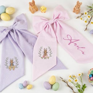 May include: Two seersucker fabric bows, one lavender and one pink, with embroidered Easter bunny designs. The pink bow has the name "Amelia" in script. Easter eggs and decorations surround the bows.