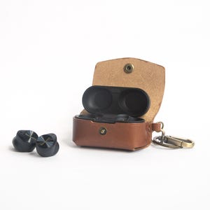 Op de afbeelding: Zwarte draadloze oordopjes met metallic accenten en merknaam. Een bruine leren etui met een drukknoopsluiting bevat de oordopjes. De etui heeft een sleutelhanger-karabijnhaak. Witte achtergrond.