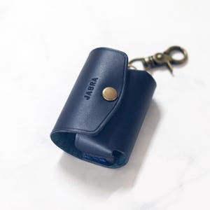 May include: A navy blue leather case with a brass-colored clasp and a snap closure. The word "JABRA" is embossed on the front. The case is designed to hold small electronics and has a key ring attachment.