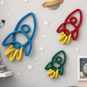 May include: A whimsical space-themed wall decor arrangement featuring a blue, red, and green rocket ship, a planet, a moon, and star accents. The room also includes framed artwork, a toy astronaut, and a wooden rocket lamp.