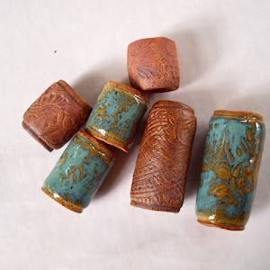 May include: A collection of handmade ceramic tumblers in various sizes and designs. Some are a reddish-brown color with carved patterns, while others are glazed in turquoise and brown. The tumblers are arranged on a white surface.