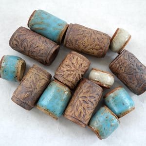 May include: A collection of handmade ceramic cups and small containers. The cups are cylindrical with a brown, textured exterior and a floral pattern. Some cups have a turquoise blue glaze on the top and bottom. The small containers are white.
