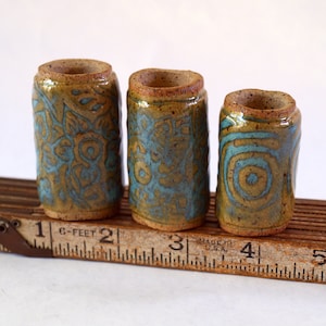 May include: Three ceramic pottery cylinders with a brown glaze and blue and green patterns. The cylinders are lined up on a wooden ruler with markings in inches.