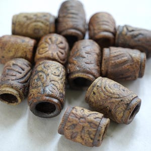 May include: A collection of cylindrical brown beads with textured surfaces featuring carved floral and geometric patterns. Each bead has a central hole and a matte, earthy finish with dark speckles, showing various intricate designs across the set.