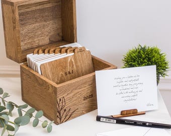 May include: A wooden box with an open lid, filled with labeled cards. The box is engraved with the initials "S&J" and the year "2025". A handwritten card and a pen are on the table. A small plant and two photos are also visible.