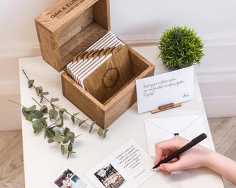 May include: A wooden box with a hinged lid, engraved with "DAN & ELISABETH AUGUST 19TH, 2023", holds several wooden cards with gold edges. A handwritten card and a pen are on the table. A small green plant and eucalyptus sprigs are also present.