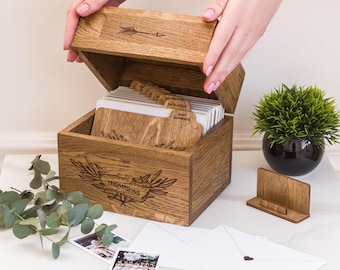 May include: Wooden keepsake box with an arrow design on the lid and the name "THOMPSONS" engraved on the front. The box is open, revealing cards with "DATE IDEAS" and "BEST MOMENTS" labels. Includes a small wooden stand and envelopes.