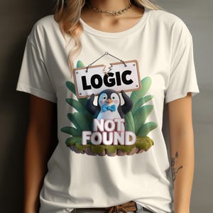 May include: Cream-colored t-shirt featuring a cartoon penguin holding a sign that reads "LOGIC NOT FOUND." The penguin has a blue bow tie and is surrounded by green leaves. The shirt is a casual wear.
