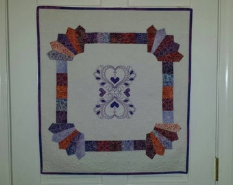 May include: A square quilt with a white center and a purple border. The quilt features a symmetrical design of hearts and fan-shaped patterns in shades of purple, orange, and blue. The quilt is hanging on a white door.