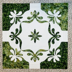 May include: A white fabric square with a green floral pattern cut out in the center. The design features a large flower in the middle and four smaller floral designs around the edges.
