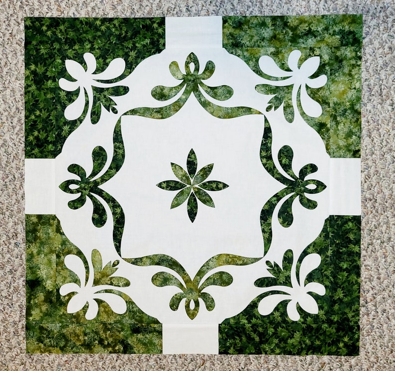 May include: A white fabric square with a green floral pattern cut out in the center. The design features a large flower in the middle and four smaller floral designs around the edges.