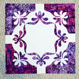 May include: A white fabric square with a decorative, cut-out design in the center. The design features a repeating pattern of stylized floral shapes. The fabric square is surrounded by four squares of purple and pink patterned fabric.