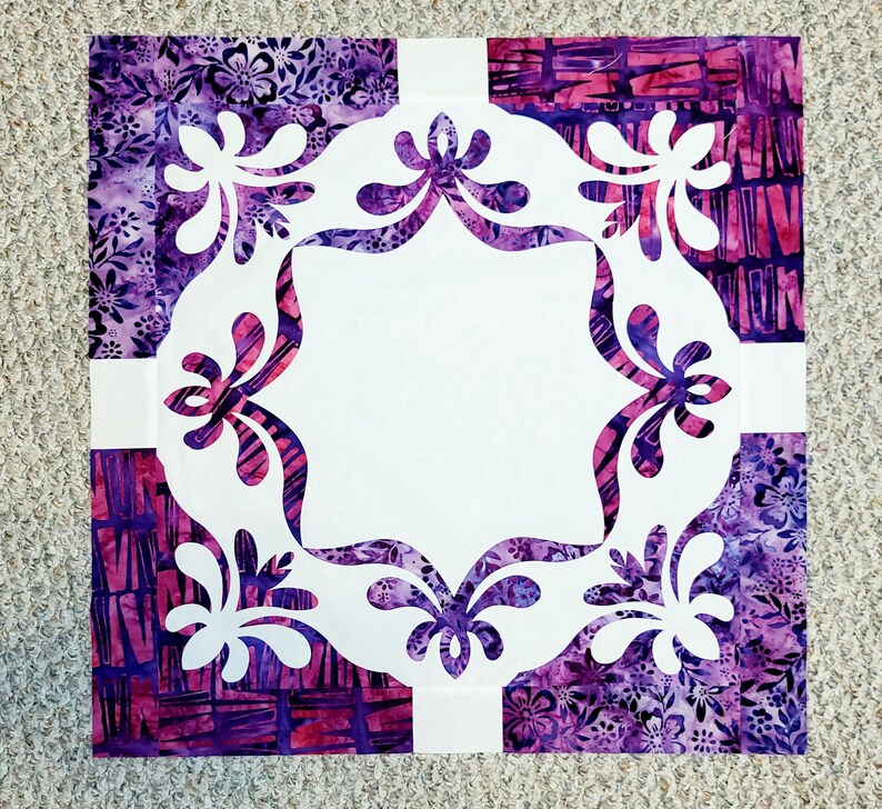 May include: A white fabric square with a decorative, cut-out design in the center. The design features a repeating pattern of stylized floral shapes. The fabric square is surrounded by four squares of purple and pink patterned fabric.