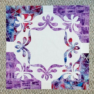 May include: A white fabric square with a decorative, white, floral design cut out of the center. The fabric square is surrounded by a purple and blue patterned fabric border.