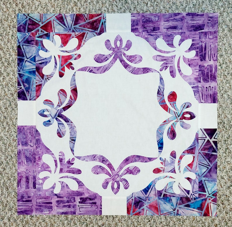 May include: A white fabric square with a decorative, white, floral design cut out of the center. The fabric square is surrounded by a purple and blue patterned fabric border.