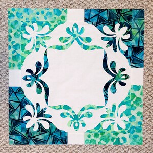 May include: A white fabric square with a decorative cut-out design of stylized flowers in the center. The fabric square is surrounded by four squares of blue and green patterned fabric.