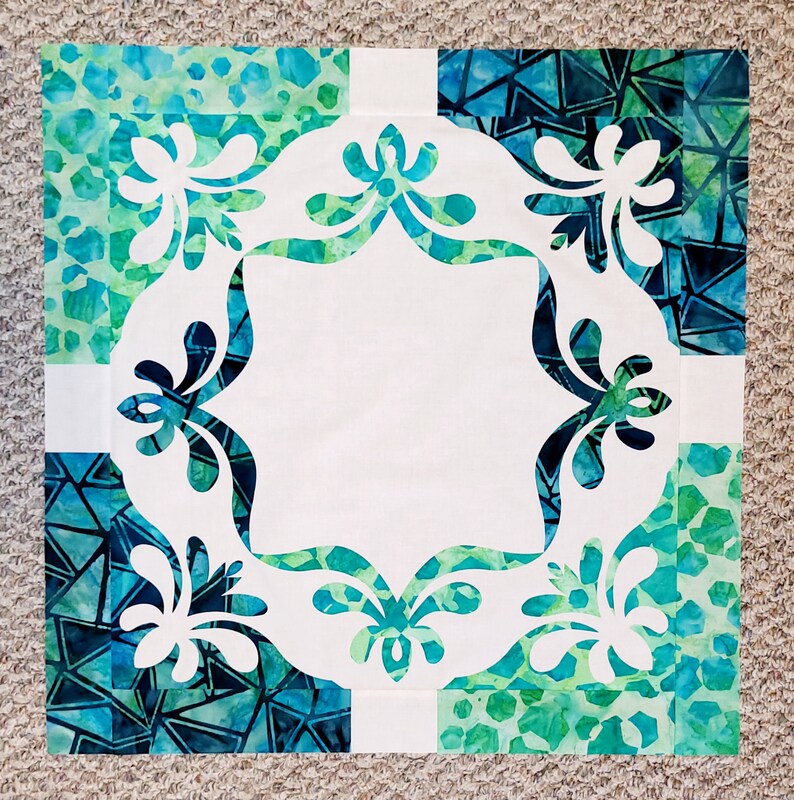May include: A white fabric square with a decorative cut-out design of stylized flowers in the center. The fabric square is surrounded by four squares of blue and green patterned fabric.