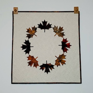 May include: A white fabric wall hanging with a brown, black, and orange maple leaf design. The leaves are arranged in a circle around a white background with a swirling pattern.