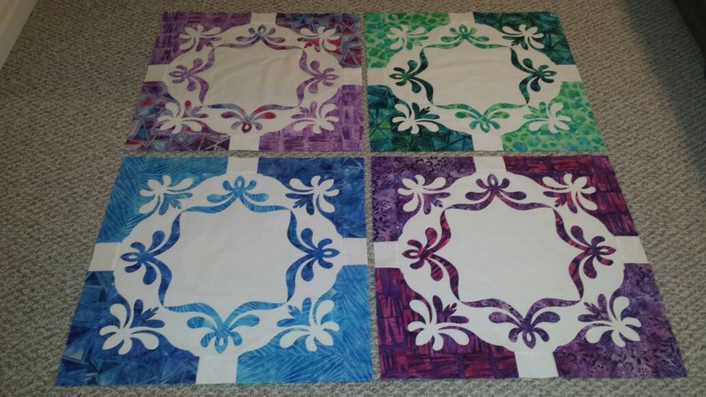 May include: Four square fabric quilt blocks with a white center and a floral design in the border. The blocks are in shades of purple, green, blue, and pink.
